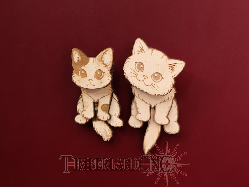 May include: Two light-colored wooden cat cutouts with engraved details, featuring large eyes and pointed ears. The left cat has a small spot on its head, while the right cat has a fluffy appearance. The text TIMBERLANDCNC is visible below.