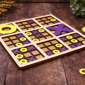 May include: A wooden tic-tac-toe game with a light brown frame, black grid, and yellow and purple game pieces. The game board is square, with the text "TIMBERLANDCNC" at the bottom. Additional yellow and purple game pieces are scattered around the board.