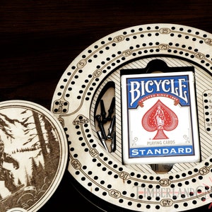 Puede incluir: Un tablero de cribbage de madera con una superficie de juego circular y una ficha de madera separada con un dise&ntilde;o de Bigfoot. El tablero tiene un compartimento para cartas y un juego de clavijas. El compartimento para cartas tiene una baraja de cartas de juego Bicycle.