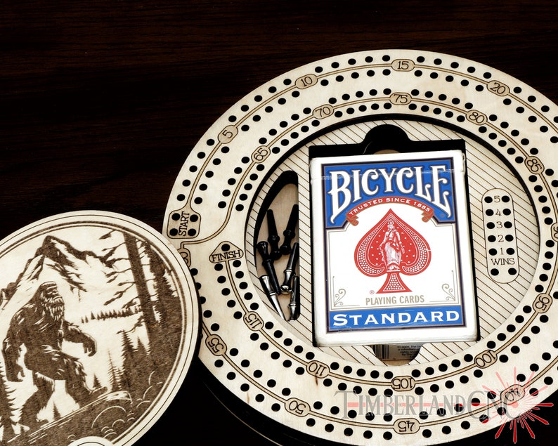 Puede incluir: Un tablero de cribbage de madera con una superficie de juego circular y una ficha de madera separada con un dise&ntilde;o de Bigfoot. El tablero tiene un compartimento para cartas y un juego de clavijas. El compartimento para cartas tiene una baraja de cartas de juego Bicycle.