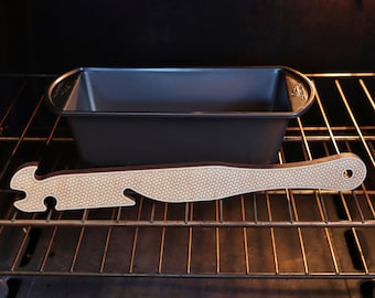 May include: A black metal loaf pan sits on a rack in an oven. A wooden spatula with a patterned design is resting on the rack.