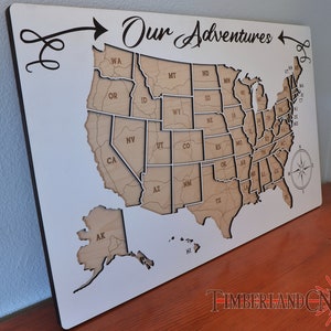 May include: A white wooden map of the United States with the words "Our Adventures" written in cursive above the map. The map is divided into individual states, each with the state abbreviation written inside. The map is mounted on a white background.