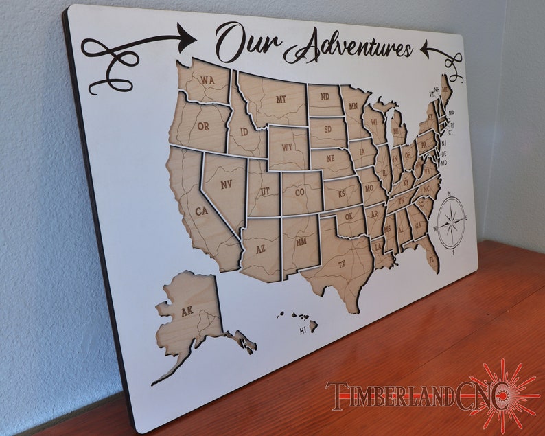 May include: A white wooden map of the United States with the words "Our Adventures" written in cursive above the map. The map is divided into individual states, each with the state abbreviation written inside. The map is mounted on a white background.