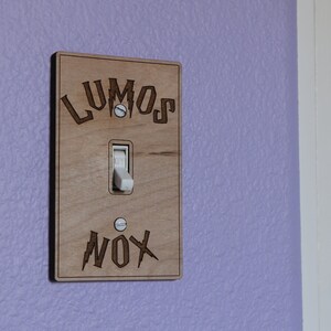 May include: A wooden light switch cover with the words "LUMOS" and "NOX" engraved on it. The switch is in the off position.