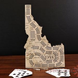May include: A wooden game board shaped like the state of Idaho with the names of cities and towns printed on it. The board has holes for pegs and is used to play a game of chance. The board is sitting on a wooden table with playing cards and pegs beside it.
