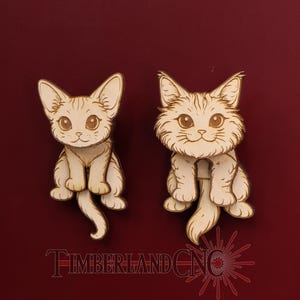 May include: Two wooden cat cutouts with detailed etched features, including large eyes and fluffy fur. The cats are light brown and sit upright with their tails curled. The text "TIMBERLAND CNC" is at the bottom of the image.