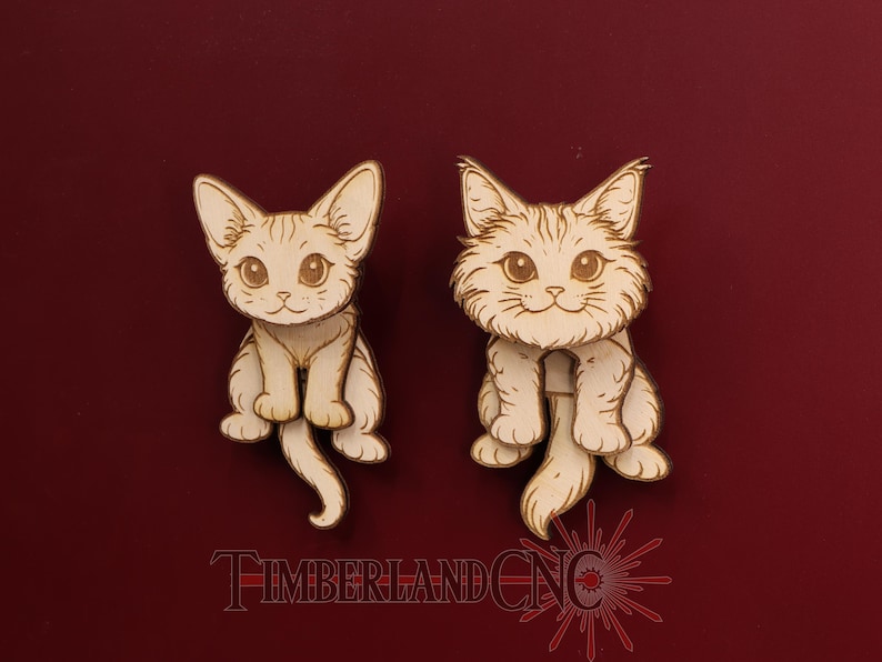 May include: Two wooden cat cutouts with detailed etched features, including large eyes and fluffy fur. The cats are light brown and sit upright with their tails curled. The text "TIMBERLAND CNC" is at the bottom of the image.