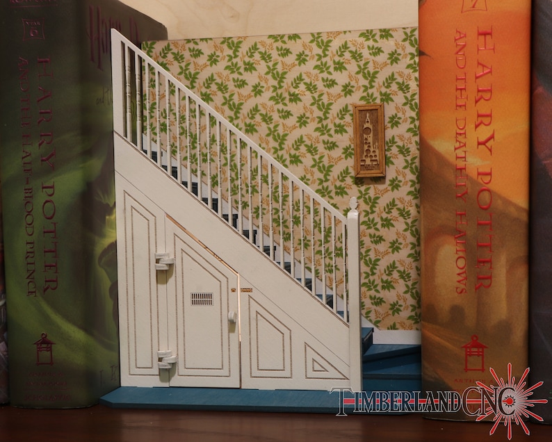 Op de afbeelding: Een witte houten trap met een witte leuning en een witte deur aan de onderkant. De trap is geplaatst tegen een groen-witte bloemenbehang. De trap maakt deel uit van een boekenleggerset, met de boeken "Harry Potter en de Relieken van de Dood" en "Harry Potter en de Halfbloed Prins" zichtbaar aan beide kanten.