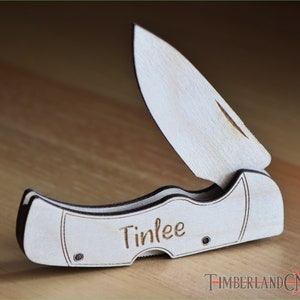 May include: A wooden folding knife with a light wood finish and dark edges. The blade is partially open, revealing a curved cutting edge. The name "Tinlee" is engraved on the handle. The knife is set against a wooden surface.