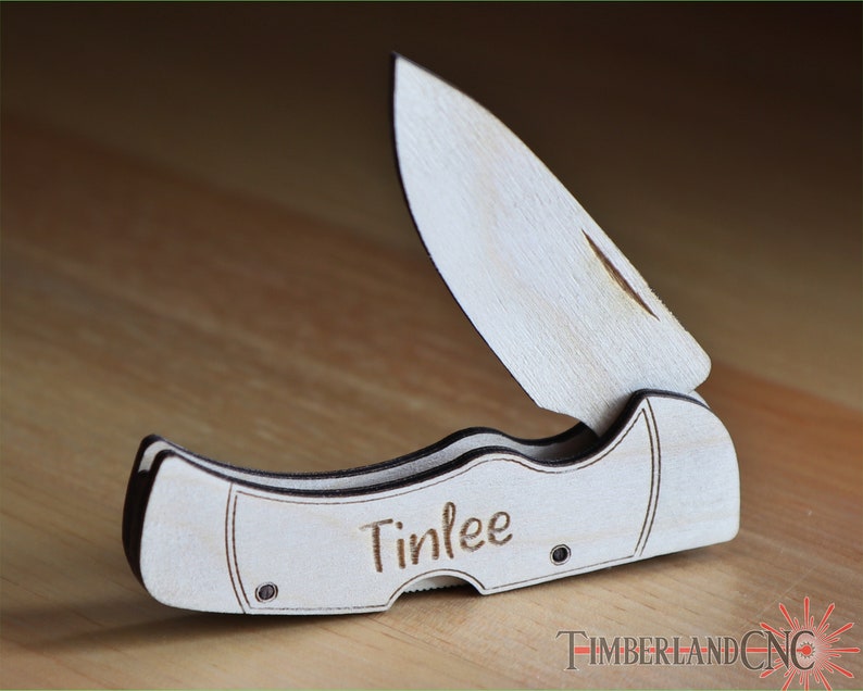May include: A wooden folding knife with a light wood finish and dark edges. The blade is partially open, revealing a curved cutting edge. The name "Tinlee" is engraved on the handle. The knife is set against a wooden surface.