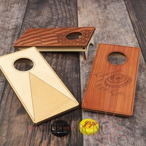 May include: Wooden cornhole game set with three boards, one featuring an American flag design. Two additional boards have circular holes and geometric patterns. Includes a black bottle cap and a yellow bottle cap.