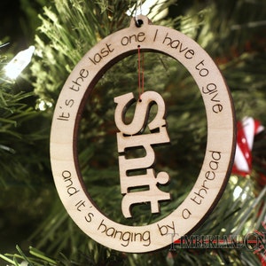 May include: A wooden Christmas ornament with the text "It's the last one I have to give and it's hanging by a thread" and the word "SHIT" cut out in the center.