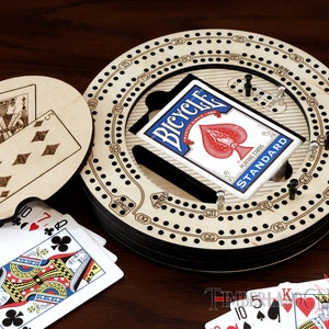 Puede incluir: Un tablero de cribbage de madera con una pista circular y una baraja de cartas Bicycle. El tablero tiene un dise&ntilde;o en blanco y negro con n&uacute;meros y puntos. Las cartas est&aacute;n en una caja negra y azul con el logotipo de Bicycle.