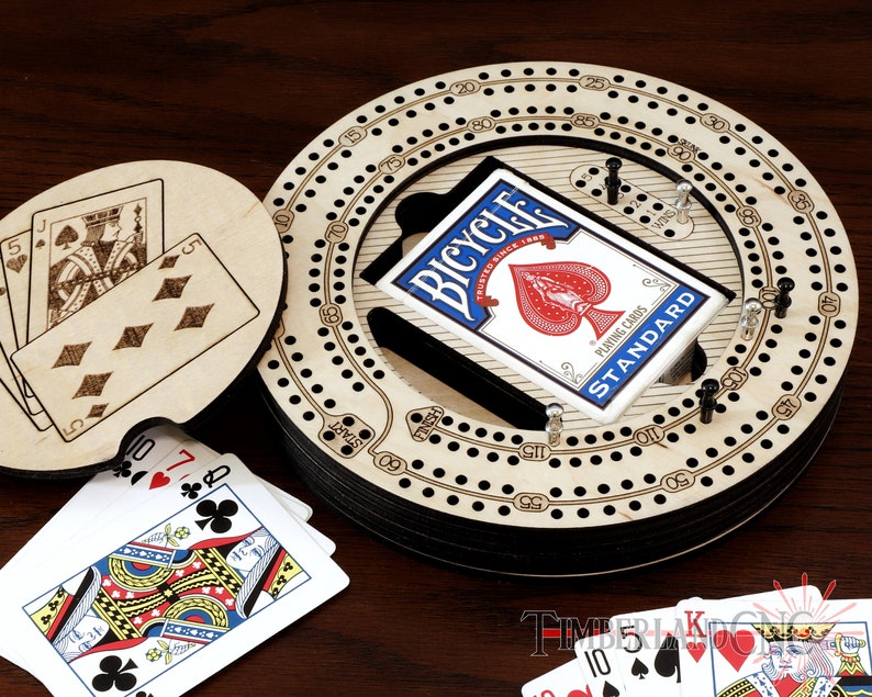 Puede incluir: Un tablero de cribbage de madera con una pista circular y una baraja de cartas Bicycle. El tablero tiene un dise&ntilde;o en blanco y negro con n&uacute;meros y puntos. Las cartas est&aacute;n en una caja negra y azul con el logotipo de Bicycle.