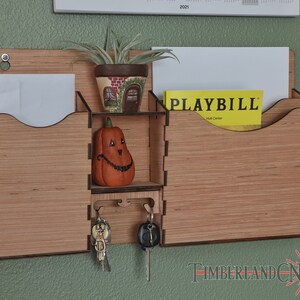 Puede incluir: Un organizador de pared de madera con dos ranuras para correo, un estante y un gancho para llaves. El organizador está hecho de madera marrón claro con una mancha marrón oscura. Hay una pequeña figura de calabaza en el estante, una planta en una maceta y una revista Playbill en una de las ranuras para correo.