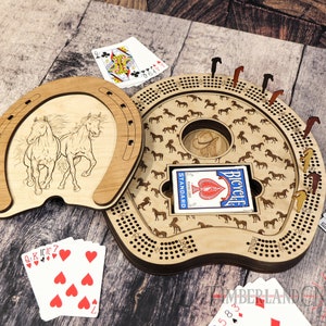 May include: A wooden horseshoe-shaped game board with a circular cutout in the centre. The board is decorated with a pattern of horses and has a small wooden horseshoe lid. The board is open and a deck of playing cards is visible inside. The board is surrounded by playing cards and wooden horses.