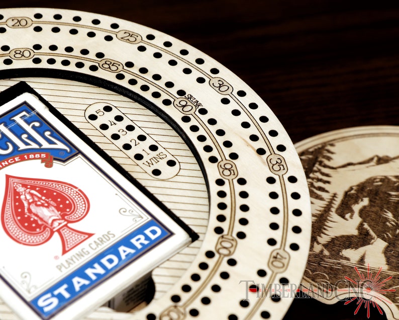Puede incluir: Un tablero de cribbage de madera con un anillo exterior negro y un c&iacute;rculo interior blanco. El tablero tiene una tarjeta de puntuaci&oacute;n con los n&uacute;meros del 1 al 5 y la palabra "WINS". Una baraja de cartas est&aacute;ndar se coloca sobre el tablero.