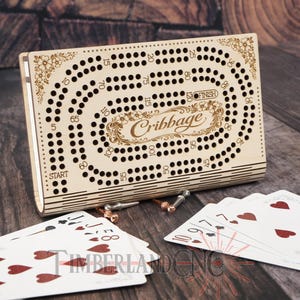 May include: A wooden cribbage board with a floral design and the word "Cribbage" engraved on the front. The board has numerous holes for pegs, with playing cards and pegs nearby. The game board is made of light-colored wood.