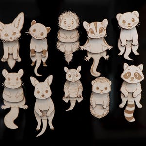 May include: A collection of ten laser-cut wooden animal magnets featuring various species including a fennec fox, hedgehog, sugar glider, armadillo, and raccoon. Each figure displays detailed engraved facial features and textures on a light wood surface.