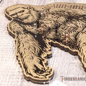May include: A wooden game board shaped like a Bigfoot with a black and brown design. The board has a grid of holes and is decorated with metal pins.