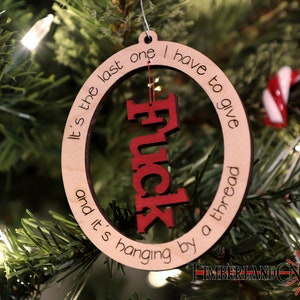 May include: A wooden Christmas ornament with a red "F*ck" cutout in the center. The ornament is round and has the text "It's the last one I have to give and it's hanging by a thread" written around the edge.