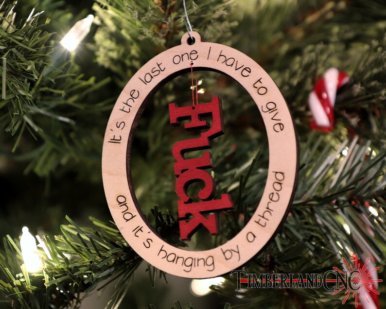 May include: A wooden Christmas ornament with a red "F*ck" cutout in the center. The ornament is round and has the text "It's the last one I have to give and it's hanging by a thread" written around the edge.