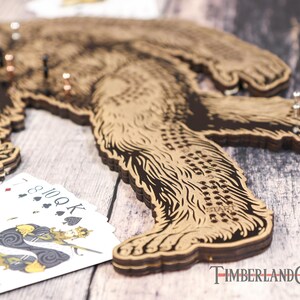 May include: A wooden Bigfoot game board with a brown and black design. The board is made of multiple layers of wood and has a textured surface. The board has a series of holes and a "START" label. Playing cards are visible in the foreground.