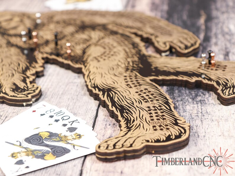 May include: A wooden Bigfoot game board with a brown and black design. The board is made of multiple layers of wood and has a textured surface. The board has a series of holes and a "START" label. Playing cards are visible in the foreground.