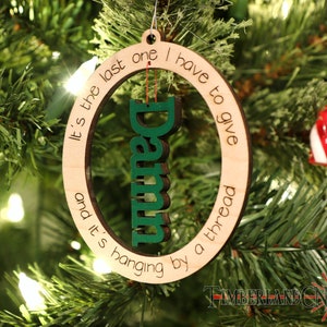 May include: A wooden Christmas ornament with the text "It's the last one I have to give and it's hanging by a thread" and the word "Damn" in green letters.