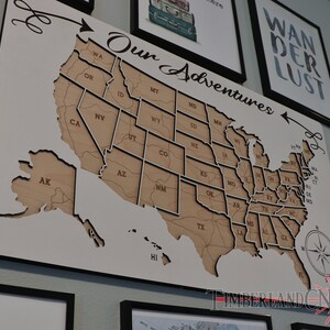 May include: A white wooden map of the United States with each state outlined. The map is mounted on a black frame and has the words "Our Adventures" written above it. The map is hanging on a wall with other framed artwork.