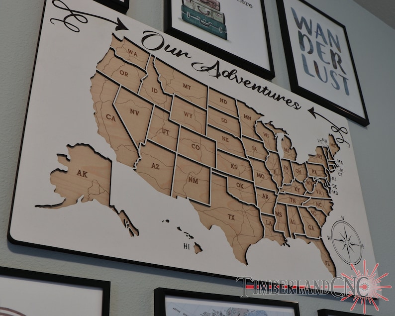 May include: A white wooden map of the United States with each state outlined. The map is mounted on a black frame and has the words "Our Adventures" written above it. The map is hanging on a wall with other framed artwork.
