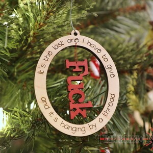 May include: A wooden Christmas ornament with the text "It's the last one I have to give and it's hanging by a thread" and the word "F*ck" in red letters inside a circle.