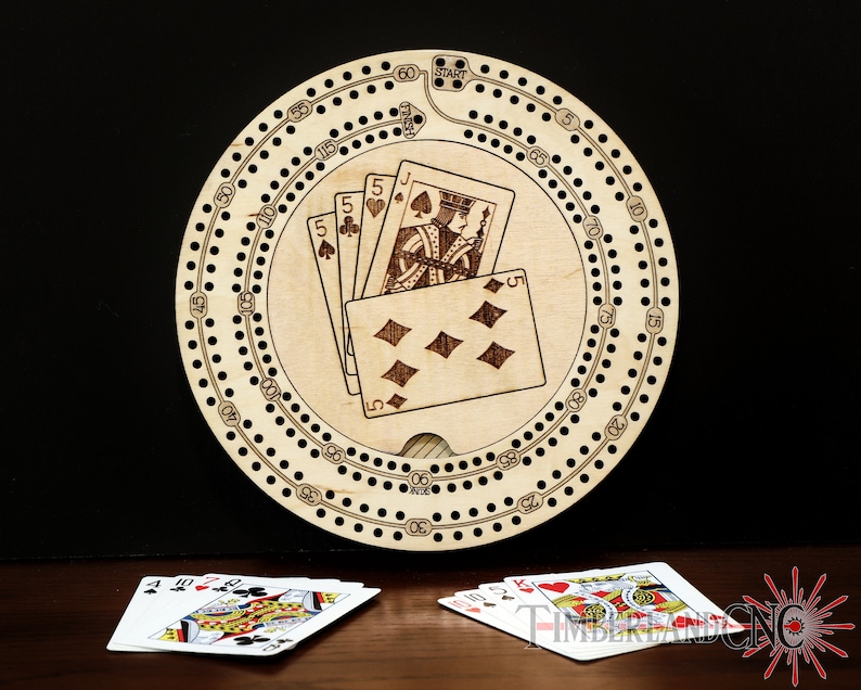 Puede incluir: Un tablero de cribbage de madera circular con una pista numerada del 1 al 60. El tablero tiene un agujero central para las fichas y una secci&oacute;n para las cartas, que muestra un 5 de picas, un 5 de tr&eacute;boles, un J de picas y un 5 de diamantes. El tablero est&aacute; sobre una mesa de madera con dos juegos de cartas.