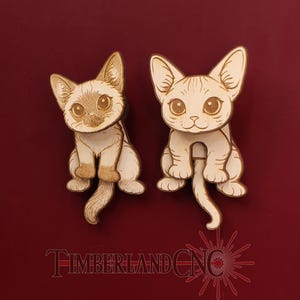 May include: Two light-colored wooden cat-shaped cutouts with engraved facial features and tails. The left cat has darker shading on its face and ears. The text TIMBERLANDCNC is printed in red at the bottom of the image.