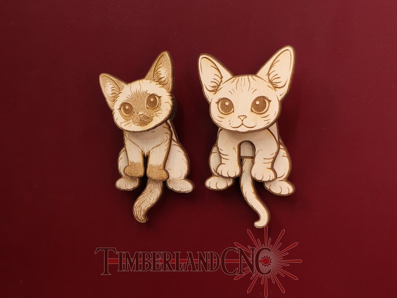 May include: Two light-colored wooden cat-shaped cutouts with engraved facial features and tails. The left cat has darker shading on its face and ears. The text TIMBERLANDCNC is printed in red at the bottom of the image.