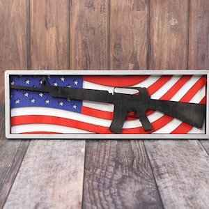 May include: A rectangular wooden wall art piece featuring a black rifle silhouette overlaid on a stylized American flag design. The flag has red and white stripes and a blue field with white stars. The artwork is set within a silver frame.