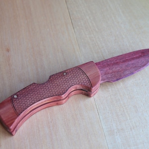 May include: A handcrafted wooden folding knife with a purple blade and a reddish-brown handle. The handle features a textured pattern and is secured with small rivets. The knife is displayed on a light-colored wooden surface.