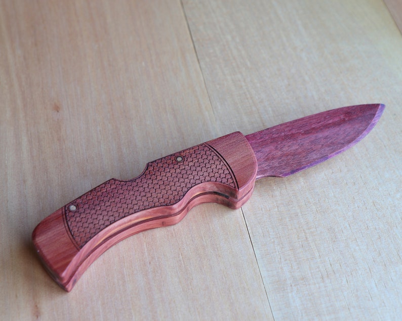 May include: A handcrafted wooden folding knife with a purple blade and a reddish-brown handle. The handle features a textured pattern and is secured with small rivets. The knife is displayed on a light-colored wooden surface.