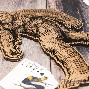 May include: A wooden Bigfoot game board with a brown and black Bigfoot design. The board has a grid of holes for playing the game. The board is on a wooden surface with playing cards and a logo that says "TimberlandCNC".
