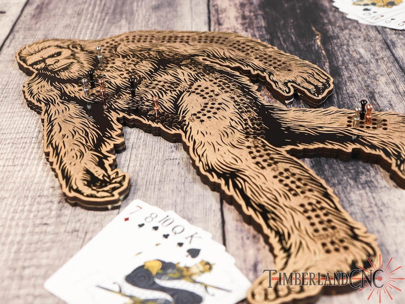 May include: A wooden Bigfoot game board with a brown and black Bigfoot design. The board has a grid of holes for playing the game. The board is on a wooden surface with playing cards and a logo that says "TimberlandCNC".