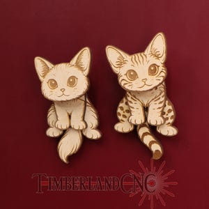 May include: Two wooden cat figurines with detailed etched designs. One cat is solid, the other has spots and stripes. Both have large eyes and are sitting upright. The text "TIMBERLAND CNC" is at the bottom.