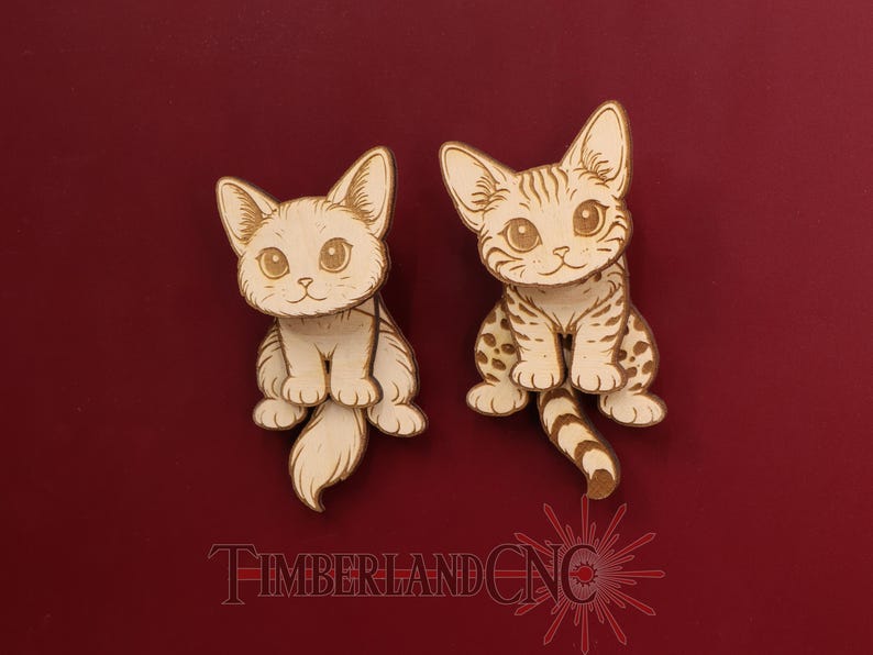 May include: Two wooden cat figurines with detailed etched designs. One cat is solid, the other has spots and stripes. Both have large eyes and are sitting upright. The text "TIMBERLAND CNC" is at the bottom.