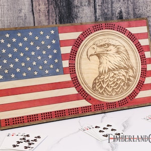 May include: A wooden cribbage board with an American flag design and a bald eagle in the center. The board has a red, white, and blue color scheme and is made of wood. The board is surrounded by a brown frame.