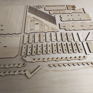 Op de afbeelding: Een set lasergesneden houten stukken voor het bouwen van een model. De stukken zijn gerangschikt op een lichtbruin oppervlak. De stukken zijn in verschillende vormen en maten, waaronder vierkanten, rechthoeken, driehoeken en trapezia.