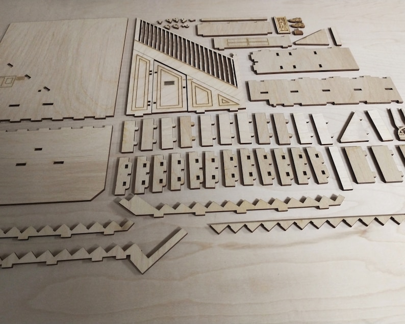 Op de afbeelding: Een set lasergesneden houten stukken voor het bouwen van een model. De stukken zijn gerangschikt op een lichtbruin oppervlak. De stukken zijn in verschillende vormen en maten, waaronder vierkanten, rechthoeken, driehoeken en trapezia.