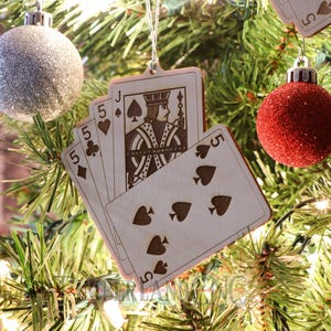 May include: Wooden playing card ornaments hanging on a Christmas tree. The ornaments feature the Jack of Spades and the number 5 on various suits. A silver glitter ball and a red glitter ball are also on the tree.