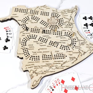 May include: A wooden game board shaped like the province of New Brunswick, Canada. The board has a grid of holes and is decorated with the names of towns and cities in the province. The board is surrounded by playing cards and game pieces.