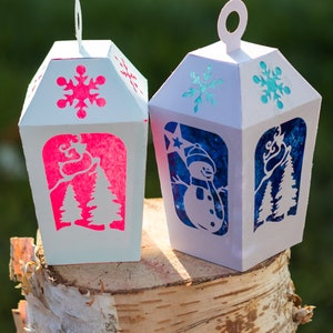 May include: Two white paper lanterns with cut-out designs. One lantern has a pink snowflake and trees, the other has a blue snowman and trees. The lanterns are sitting on a birch log.