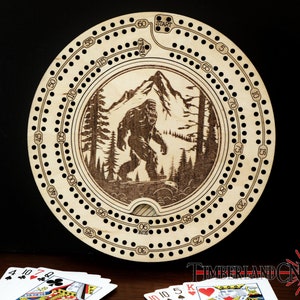 Puede incluir: Un tablero de cribbage de madera con un dise&ntilde;o grabado con l&aacute;ser de Bigfoot caminando por un bosque. El tablero tiene una forma circular con un agujero central y una pista de agujeros numerados alrededor del borde. El tablero est&aacute; hecho de madera de color claro y tiene un acabado natural.