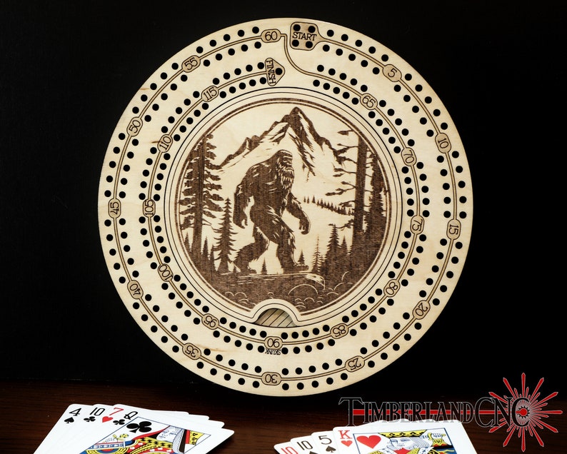 Puede incluir: Un tablero de cribbage de madera con un dise&ntilde;o grabado con l&aacute;ser de Bigfoot caminando por un bosque. El tablero tiene una forma circular con un agujero central y una pista de agujeros numerados alrededor del borde. El tablero est&aacute; hecho de madera de color claro y tiene un acabado natural.